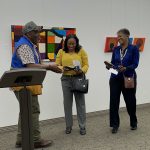 Three people standing in front of art in a gallery and one person standing at a podium