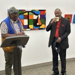 Two men standing in front of art in a gallery. One man behind a podium.