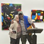 Two people standing and hugging in front of art in a gallery. One man standing in front of a podium.