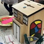 Ballot box shaped and painted like a house with windows and doors. Ballot slips in front of the box.