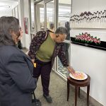 One person admiring art on a wall in an art gallery. One person grabbing a piece of candy from a table below the art on the wall.