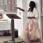 Woman in a shirt and skirt leaning against the wall behind the podium pointing out in front of her.