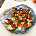 A plate of snacks that look like ladybugs made up of crackers, black olive, tomato, cucumber, and a sauce.