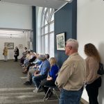 People sitting and standing along a wall looking towards the left.