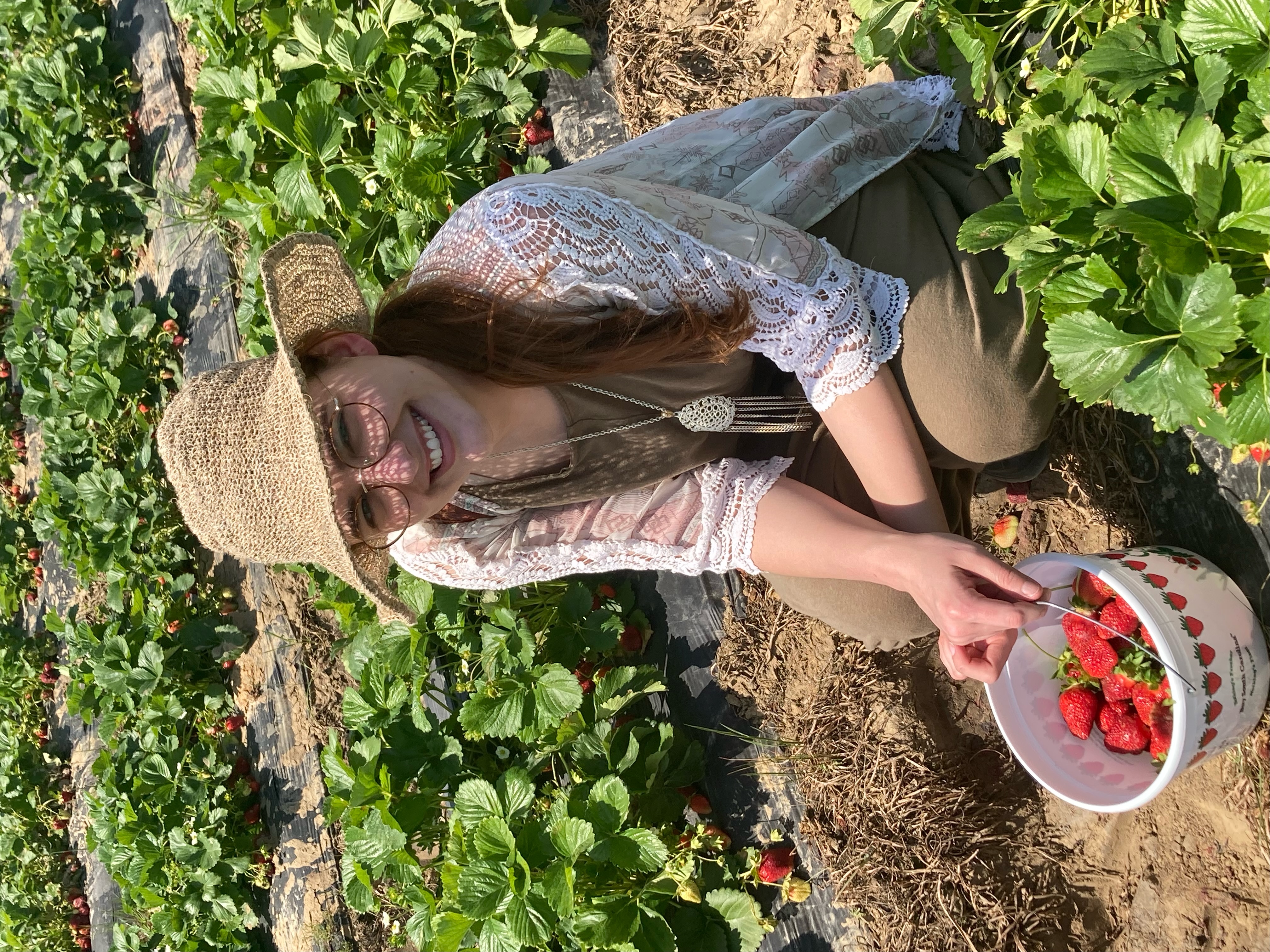Mikaela crouched down in a strawberry field with a bucket of strawberries