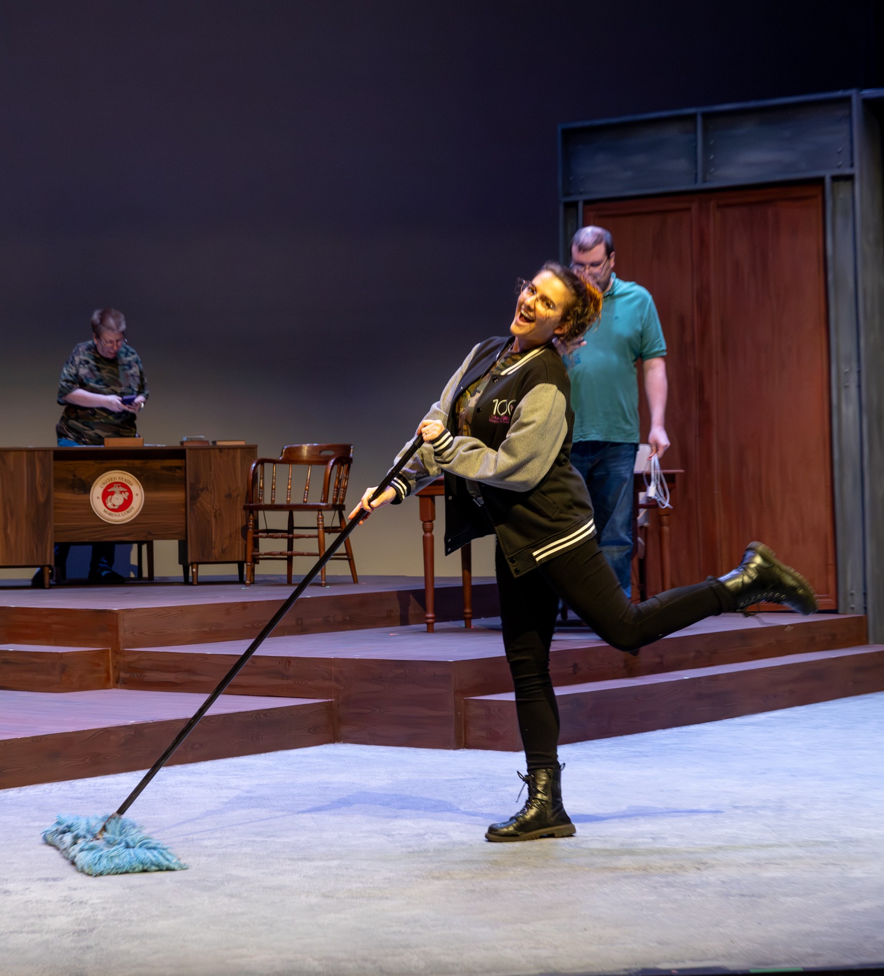 Mikaela is pushing a broom across a theater set while smiling and kicking up her foot