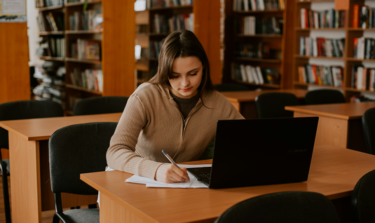 Teen girl sitting in library with open laptop and pencil in hand